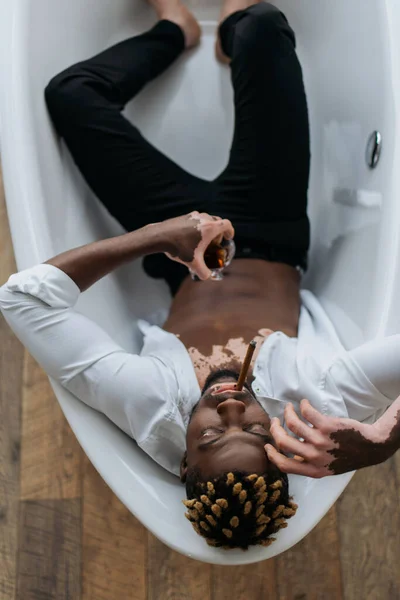 Visão aérea do homem afro-americano com vitiligo na roupa fumando charuto e segurando uísque na banheira — Fotografia de Stock
