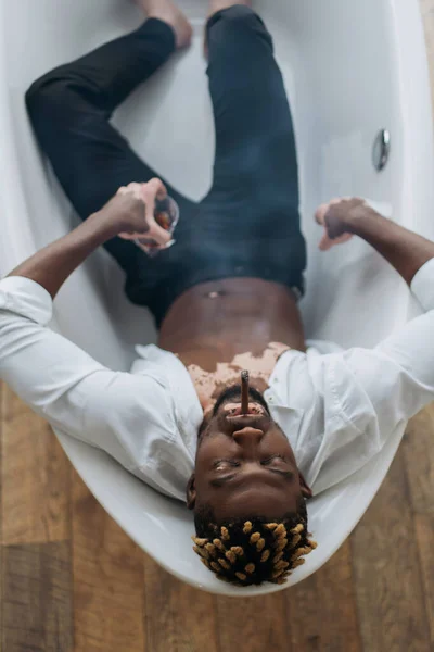 Vista aérea del hombre afroamericano con vitiligo en camisa y pantalones fumando cigarro y sosteniendo whisky en la bañera - foto de stock