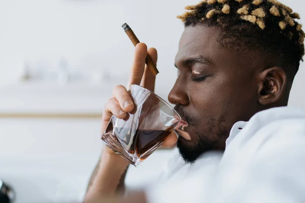 Бічний вид на афроамериканського чоловіка з вітиліго, який тримає сигару і п'є віскі у ванній кімнаті. — стокове фото