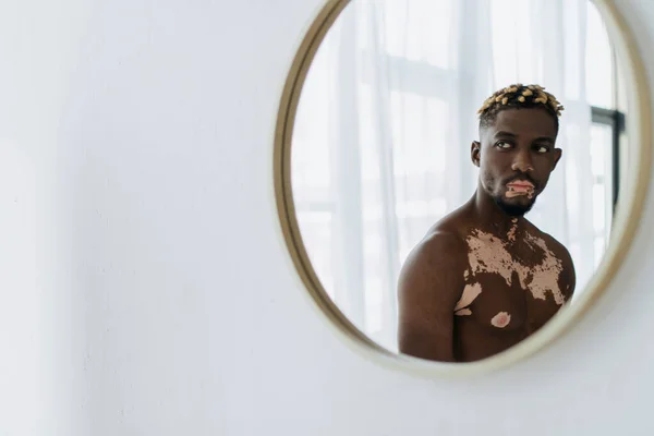 Hombre afroamericano sin camisa con vitiligo que se refleja en el espejo de baño moderno - foto de stock