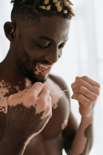 Улыбающийся и без рубашки африканский американец с витилиго, держащий зубную нить дома — стоковое фото