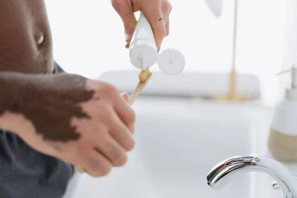 Vista recortada del hombre afroamericano con vitiligo apretando pasta de dientes en el cepillo de dientes en el baño - foto de stock