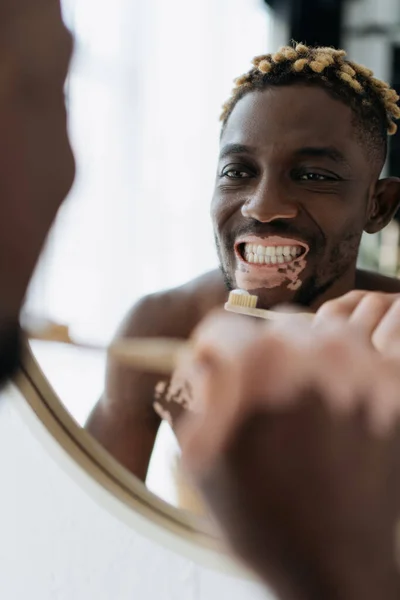 Africano americano com vitiligo olhando para os dentes e segurando escova de dentes perto do espelho em casa — Fotografia de Stock