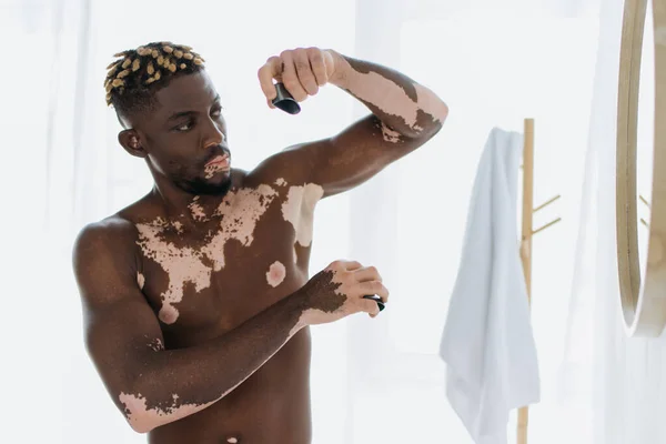 Uomo africano americano senza camicia con vitiligine utilizzando deodorante vicino specchio in bagno — Foto stock