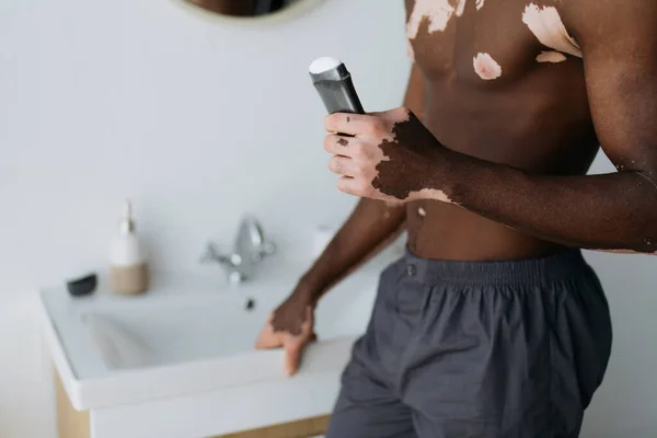 Cropped view of muscular african american man with vitiligo holding antiperspirant in bathroom — Stock Photo