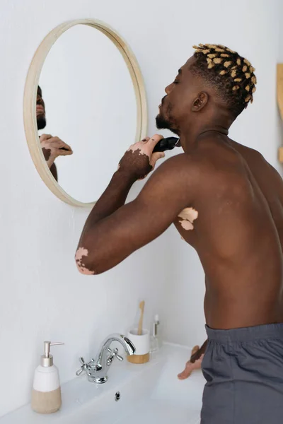Vista lateral del hombre afroamericano con vitiligo afeitado con afeitadora eléctrica en el baño - foto de stock