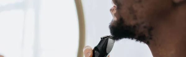 Cropped view of african american man with vitiligo shaving beard with electric razor in bathroom, banner — Stock Photo