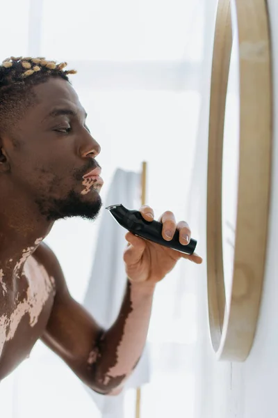 Африканский мужчина без рубашки с витилиго с электрической бритвой возле зеркала в ванной комнате — стоковое фото