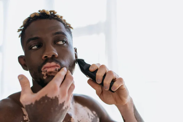 African american man with vitiligo shaving beard with electric razor at home in morning — Stock Photo