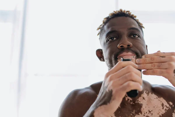 Мускулистый африканский американец с витилиго бритья с электрической бритвой в ванной комнате — стоковое фото