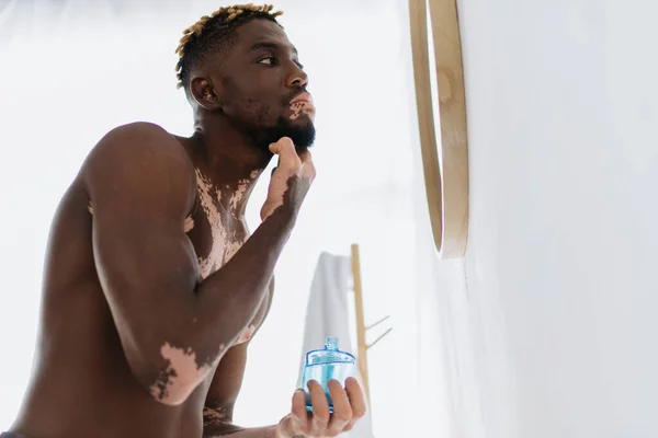 Vista basso angolo di uomo africano americano senza camicia con vitiligine applicazione lozione sul viso in bagno — Foto stock