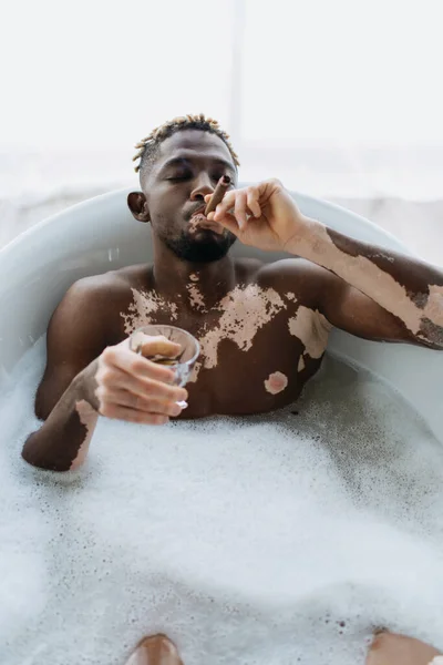 African american man with vitiligo smoking cigar and holding whiskey while taking bath at home — Stock Photo
