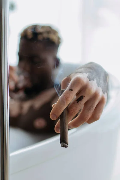 Blurred africano americano hombre con vitiligo celebración cigarro mientras toma baño en casa - foto de stock