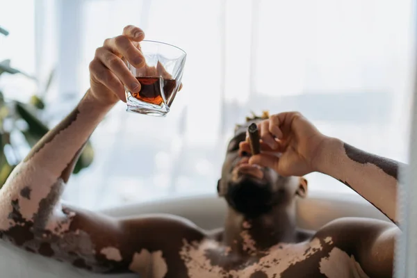 Homem americano africano desfocado com vitiligo fumando charuto e segurando uísque enquanto toma banho em casa — Fotografia de Stock