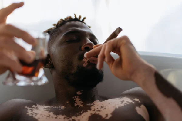 Young african american man with vitiligo smoking cigar and holding blurred whiskey while taking bath at home — Stock Photo