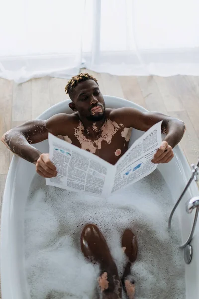 Vista superior do homem americano africano com vitiligo ler jornal enquanto toma banho em casa — Fotografia de Stock