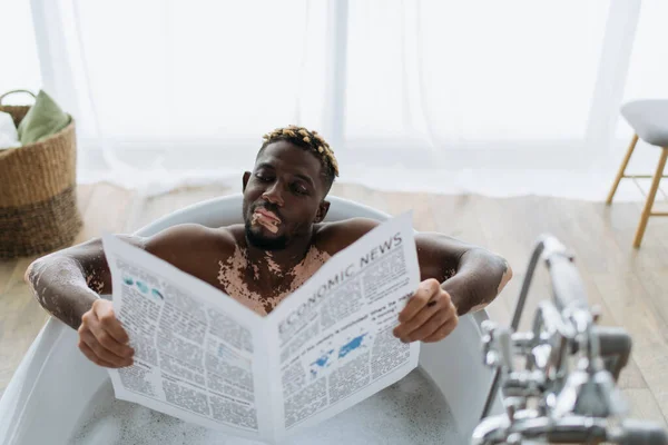 Joven afroamericano con vitiligo leyendo periódico económico mientras toma baño en casa - foto de stock