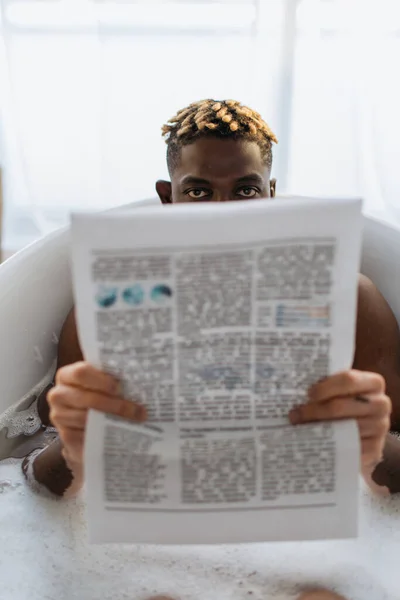 African american man with vitiligo covering face with newspaper in bath with foam at home — Stock Photo