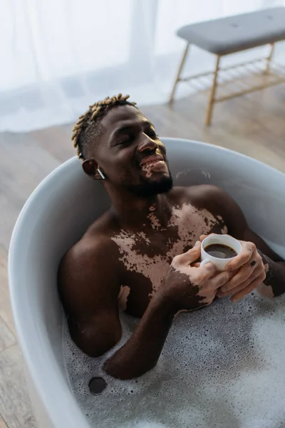 High angle view of smiling african american man with vitiligo using earphone and holding coffee in bathtub — Stock Photo