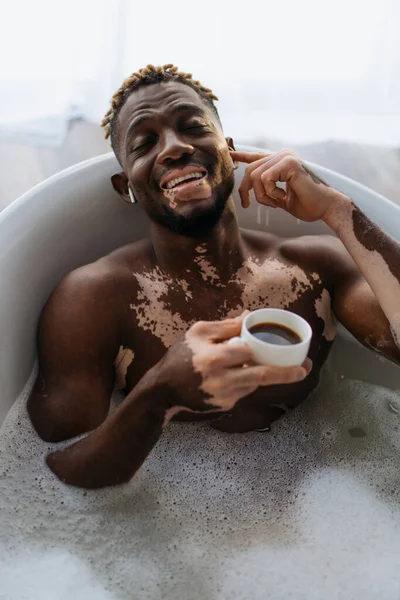 Heureux homme afro-américain avec vitiligo en utilisant des écouteurs et en tenant une tasse de café tout en prenant un bain à la maison — Photo de stock