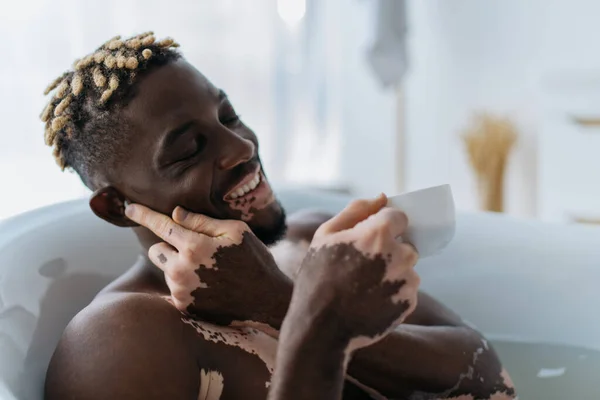 Homem afro-americano muito feliz com vitiligo usando fone de ouvido sem fio e segurando xícara de café na banheira em casa — Fotografia de Stock