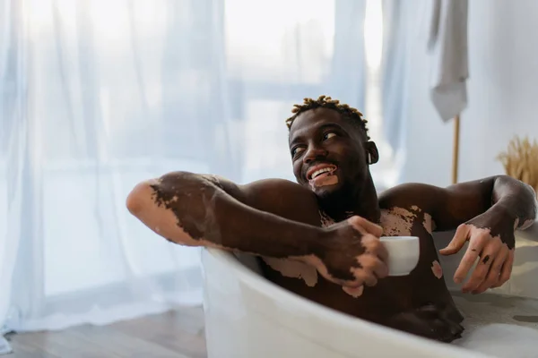 Веселый африканский американец с витилиго в наушниках держа чашку кофе и принимая ванну дома — стоковое фото