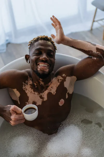 Top view of positive african american man with vitiligo listening music in earphone and holding coffee cup in bathtub with foam — Stock Photo