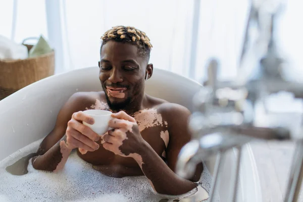 Sonriente hombre afroamericano con vitiligo escuchando música en auriculares y sosteniendo café mientras toma un baño - foto de stock