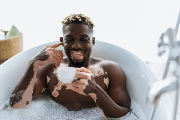 Sorridente uomo africano americano con la vitiligine ascoltare musica in auricolare e tenere in mano la tazza di caffè mentre si fa il bagno a casa — Foto stock