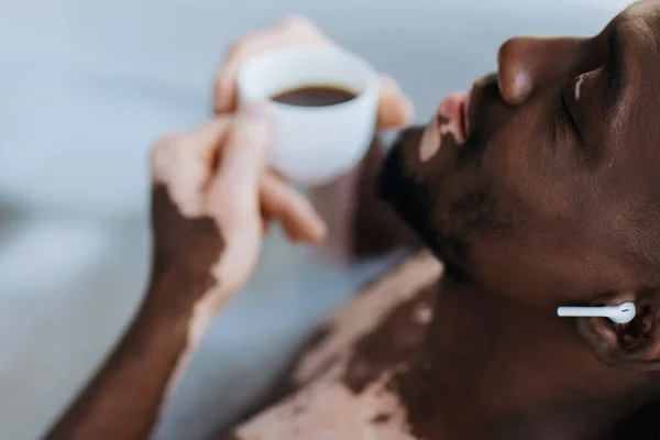 Afrikanischer Mann mit Vitiligo mit Kopfhörer und hält verschwommenen Kaffee, während er zu Hause baden geht — Stockfoto