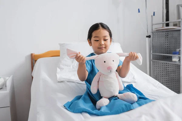 Carefree asian girl sitting on bed in hospital and playing with toy bunny — Stock Photo