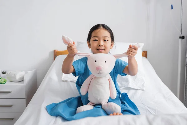 Asian girl in hospital gown smiling at camera while playing with toy bunny on bed in clinic — Stock Photo