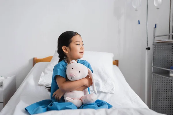 Стомлена азіатська дівчина обіймає іграшкового кролика і дивиться геть, сидячи на лікарняному ліжку — стокове фото