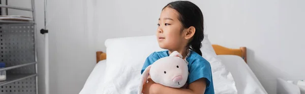 Dreamy asian child embracing toy bunny and looking away in hospital ward, banner — Stock Photo