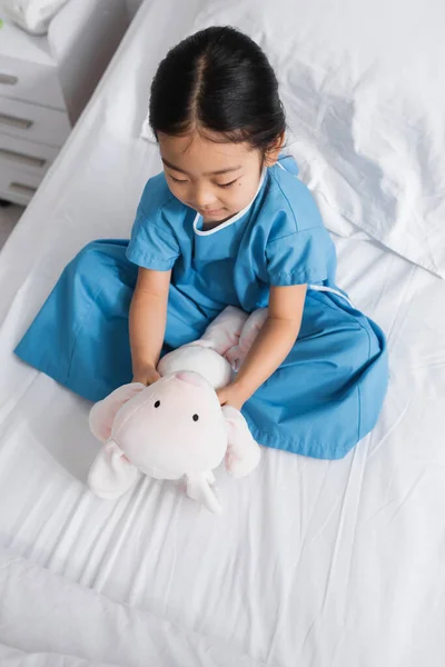 Top view of little asian girl sitting on bed and playing with toy bunny in hospital ward — Stock Photo