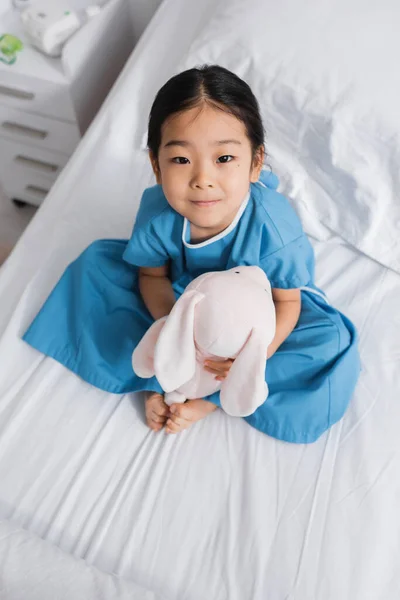 Draufsicht auf kleine asiatische Mädchen sitzt auf Krankenhausbett mit Spielzeughase und schaut in die Kamera — Stockfoto