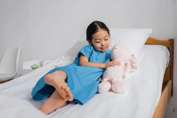 Босоногая азиатка в больничном платье, обнимающая игрушечного кролика, лежащего на кровати в педиатрической клинике — стоковое фото