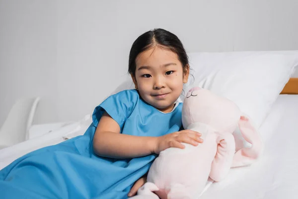 Довольная азиатская девушка обнимает игрушечного кролика и смотрит в камеру на кровати в больничном отделении — стоковое фото