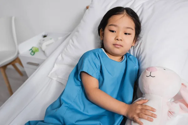Pensativo asiático niño acostado en hospital cama con juguete conejito y mirando lejos - foto de stock