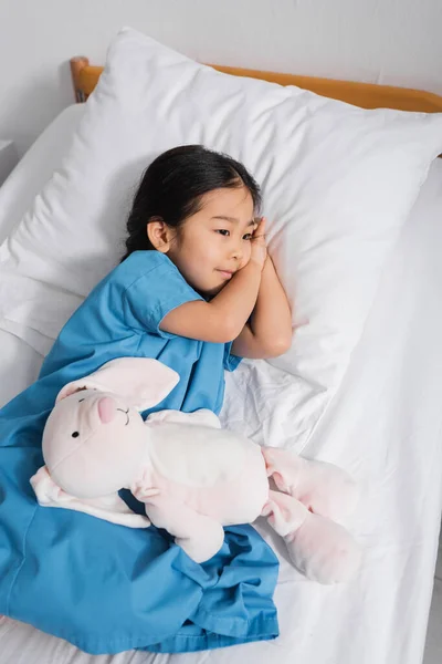 Високий кут зору мрійлива азіатська дитина лежить на лікарняному ліжку біля іграшкового кролика і дивиться в сторону — стокове фото