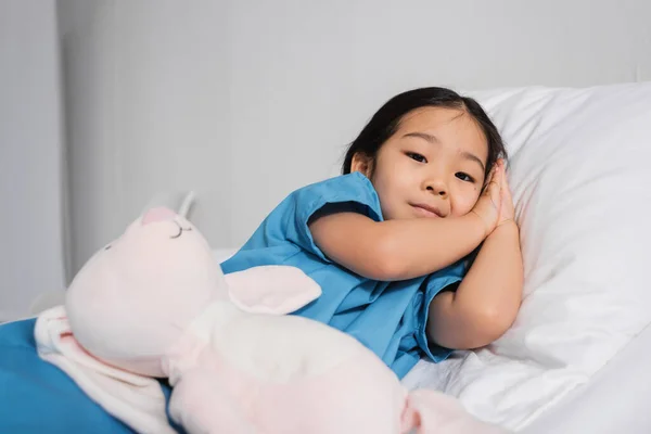 Позитивна азіатська дитина дивиться на камеру, лежачи на лікарняному ліжку біля іграшкового кролика — стокове фото