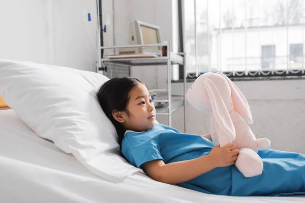 Вид збоку азіатської дівчини в лікарняній сукні, дивлячись на іграшкового кролика, лежачи на ліжку в клініці — стокове фото
