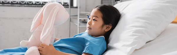 Вид сбоку азиатского ребенка, смотрящего на игрушечного кролика, лежащего на больничной койке в клинике, баннер — стоковое фото