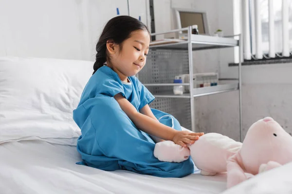 Sonriente asiático chica en hospital bata jugando con juguete conejito en cama en pediátrico clínica - foto de stock
