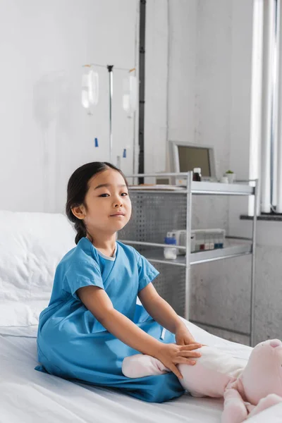 Asiático chica en hospital bata sentado en cama cerca juguete conejito y mirando cámara - foto de stock