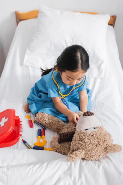 Високий кут зору азіатської дівчини, яка вивчає плюшевого ведмедя з іграшковим стетоскопом під час гри на ліжку в лікарні — стокове фото