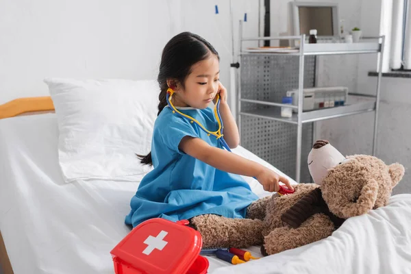 Pouco ásia menina examinando ursinho de pelúcia com brinquedo estetoscópio no hospital cama — Fotografia de Stock