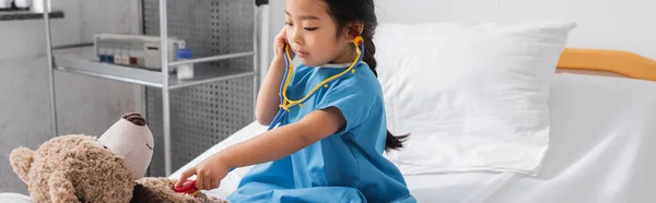 Asian girl examining teddy bear with toy stethoscope on hospital bed, banner — Stock Photo