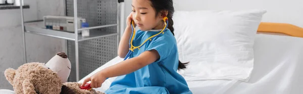 Asiático chica examinar teddy oso con juguete estetoscopio en hospital cama, banner - foto de stock