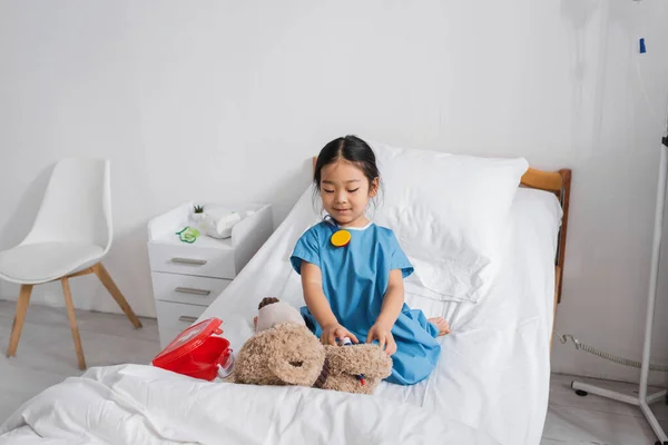 Веселый азиатский ребенок играет с плюшевым мишкой и игрушечным медицинским оборудованием на кровати в педиатрической клинике — стоковое фото